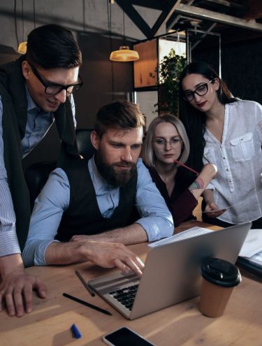 two men and two women gathered, leaning forward and looking in one direction, possibly toward a computer monitor
