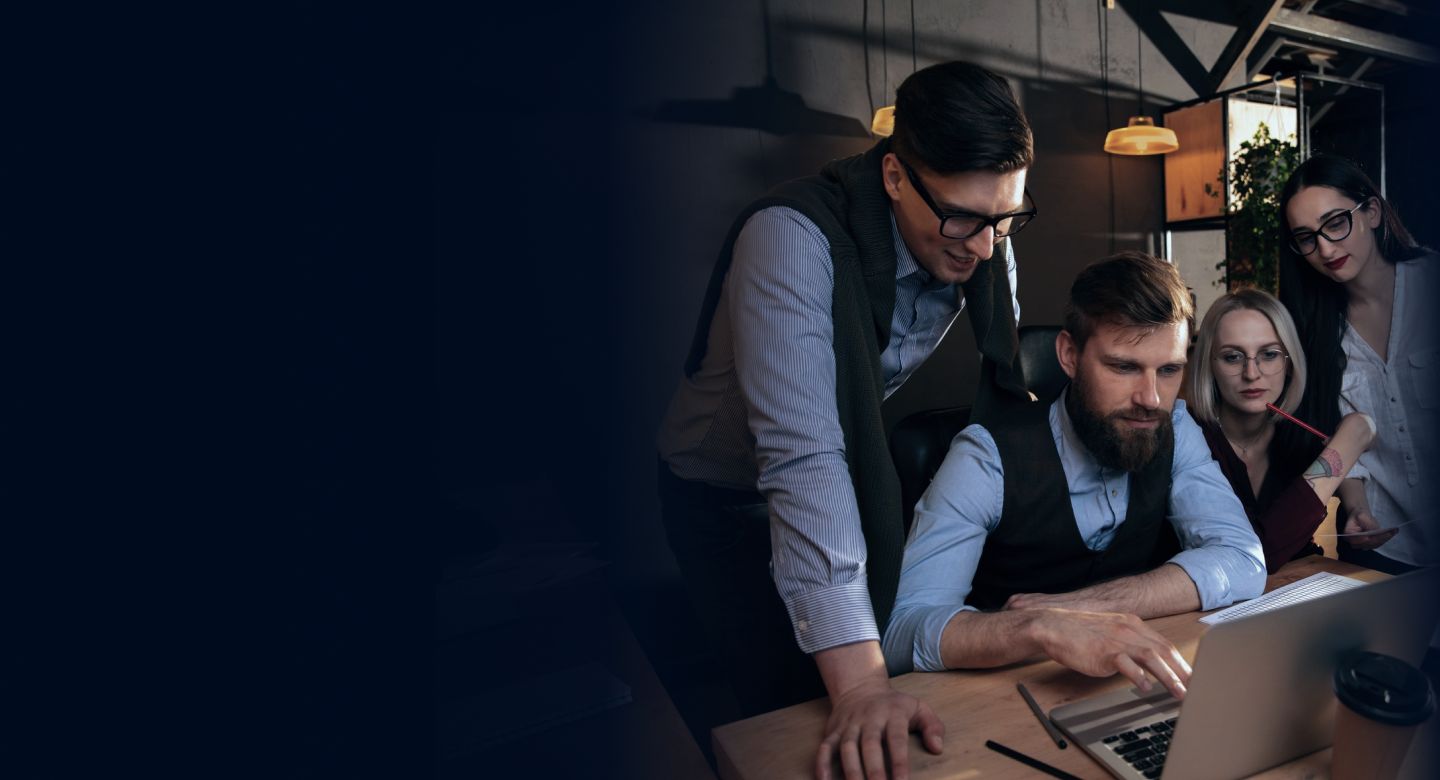 two men and two women gathered, leaning forward and looking in one direction, possibly toward a computer monitor